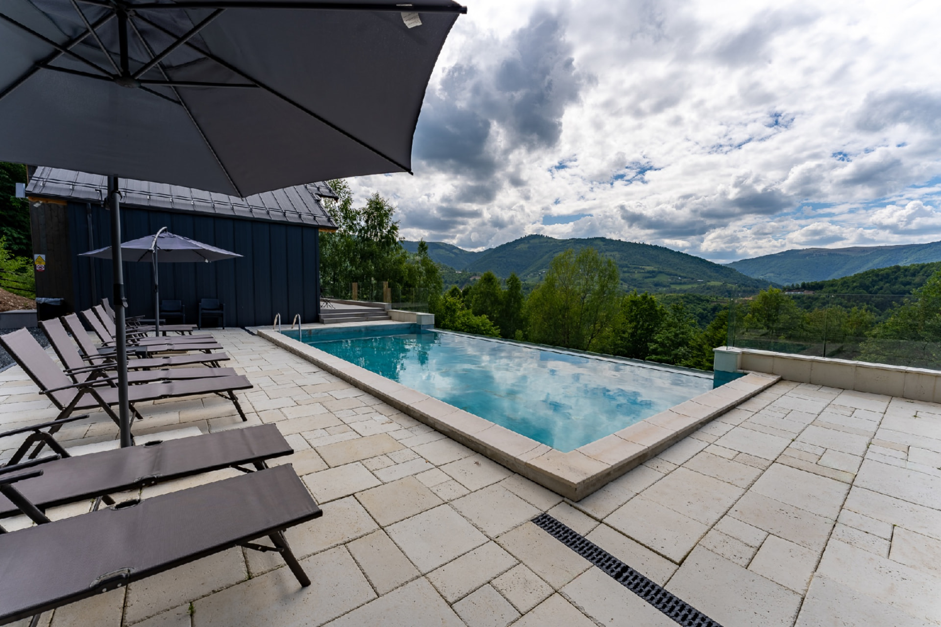 Terasa cu piscină, umbrele și șezlonguri lângă cabana cu fațadă închisă, deasupra văii cu dealuri verzi